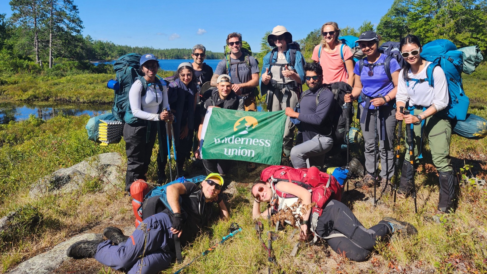 Thirteen people in full backpacking packs grouped on a rocky, grassy point beside a lake under a clear blue sky, two of them lying in the foreground grass, holding a green Wilderness Union flag.